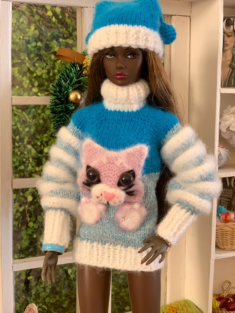 Pu&ograve; includere: Una bambola che indossa un maglione di lana blu e bianco con un design di gatto rosa. La bambola indossa anche un cappello di lana blu e bianco.