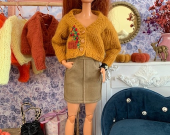 Può includere: Una bambola con lunghi capelli rossi indossa un cardigan in maglia giallo senape con un piccolo motivo floreale ricamato e tre bottoni, abbinato a una minigonna in velluto a coste color marrone chiaro. La bambola ha orecchini verdi e scarpe nere.