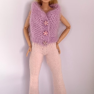 Peut inclure: Un chapeau en tricot rose, un gilet en tricot violet avec des boutons en forme de fleurs roses et un pantalon en tricot rose sur une poupée.