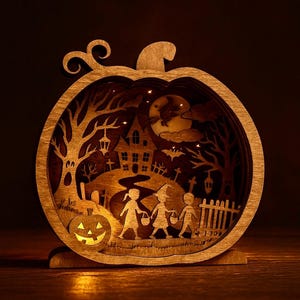 Könnte beinhalten: Holz-Halloween-Dekoration in Kürbisform. Das geschichtete Design zeigt eine gruselige Szene mit einem Spukhaus, Bäumen, Kindern, die Süßes oder Saures machen, und einer Jack-o'-Lantern. Der Kürbis hat ein Innenlicht, das die Szene beleuchtet.