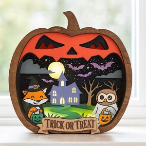 Op de afbeelding: Houten Halloween decoratie in de vorm van een pompoen. De pompoen heeft een uitgesneden gezicht en bevat een gelaagde scène met een vos, een uil, een spookhuis, vleermuizen en de tekst "Trick or Treat".