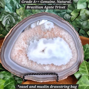 May include: A Brazilian agate trivet with a natural, layered design in shades of white, gray, and brown. The trivet is displayed on a black metal stand. Text on the image reads: "Grade A++ Genuine, Natural, Brazilian Agate Trivet" and "*easel and muslin drawstring bag included*"