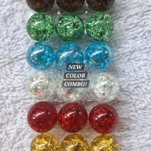 May include: A collection of eighteen spherical glass beads arranged in six rows of three, featuring a crackled texture in brown, green, blue, clear, red, and yellow. The center of the display includes a black rectangular label with the white text NEW COLOR COMBO!