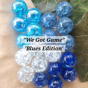 May include: A collection of round crackle glass marbles in various shades of blue, light blue, and clear, arranged in a cluster. The text "We Got Game" and "Blues Edition" is overlaid in the center of the marbles in a white rectangular box.