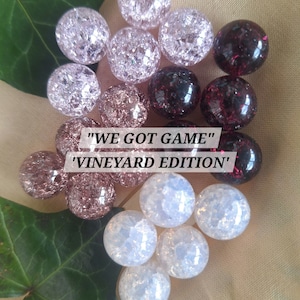 May include: A collection of round, crackle-textured glass beads in shades of white, light pink, and dark burgundy. The text "WE GOT GAME" and "VINEYARD EDITION" is centered over the beads. The beads are arranged alongside green leaves.