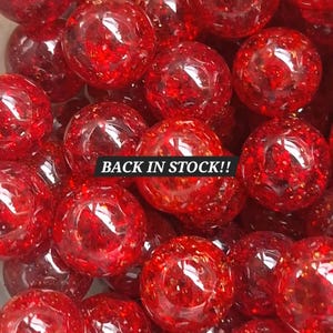 May include: A close-up of a pile of red glass marbles. The marbles are transparent with a speckled, glittery appearance. The text "BACK IN STOCK!!" is displayed across the image in a white font.