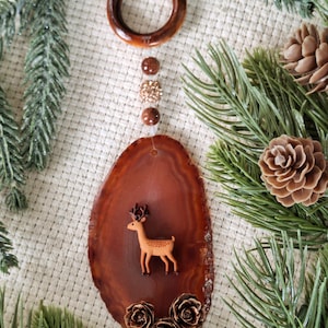 May include: A brown agate slice Christmas tree ornament with a small deer figurine and three brown pinecones. The ornament is hanging from a brown wooden ring with gold and brown beads.