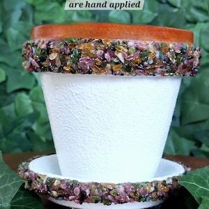 Puede incluir: Maceta de cerámica blanca con borde marrón y una banda de piedras preciosas de turmalina de colores alrededor de la parte superior. La maceta tiene un platillo a juego con una banda de piedras preciosas. El texto "All genuine tourmaline gemstones are hand applied" está escrito sobre un fondo verde encima de la maceta.