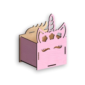 Peut inclure: Une boîte en bois rose en forme de tête de licorne. La boîte a un motif découpé d'un visage de licorne avec les yeux fermés, des cils et une corne. La boîte a un couvercle en bois avec une fente pour insérer des objets.