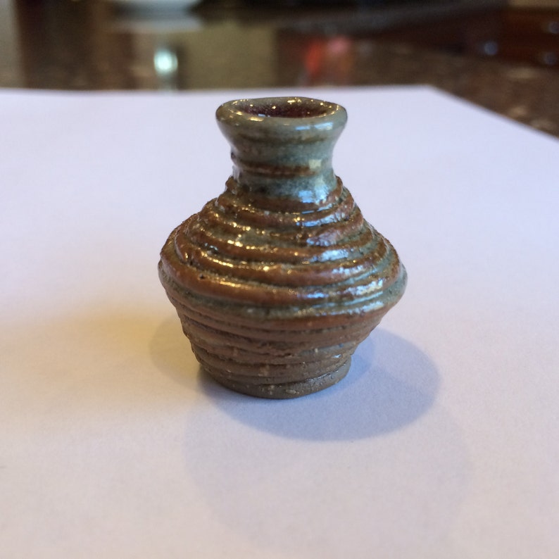 May include: A small, brown and gray ceramic vase with a rounded body and a narrow neck. The vase is decorated with a series of horizontal lines.