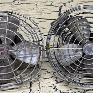Puede incluir: Dos ventiladores de metal vintage con un diseño de jaula circular. Los ventiladores tienen tres aspas y un cubo central con un emblema rojo. El metal tiene una apariencia desgastada y envejecida, lo que sugiere que son artículos antiguos o retro.