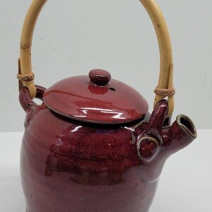 May include: A burgundy ceramic teapot with a bamboo handle. The teapot has a lid with a small knob and a spout. The handle is arched and attached to the body with woven twine. The teapot is a deep red color.