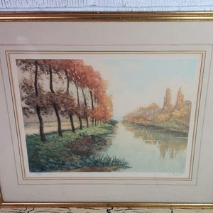 May include: A framed landscape print featuring a river scene with trees lining the banks. The trees display autumnal colors, with reds and browns dominating. The artwork is set within a gold-colored frame.