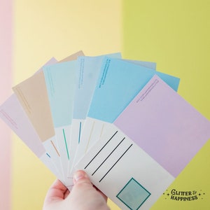 Pode incluir: Um conjunto de seis cart&otilde;es postais em branco em v&aacute;rias cores pastel, incluindo azul claro, rosa, amarelo e bege. Os cart&otilde;es postais t&ecirc;m um espa&ccedil;o em branco para escrever e uma &aacute;rea para o selo. A parte de tr&aacute;s dos cart&otilde;es postais tem uma linha para o endere&ccedil;o e uma linha para o endere&ccedil;o do remetente. Os cart&otilde;es postais est&atilde;o dispostos em forma de leque e s&atilde;o mantidos em uma m&atilde;o. O texto "Glitter & Happiness" est&aacute; impresso no canto inferior direito do cart&atilde;o postal.