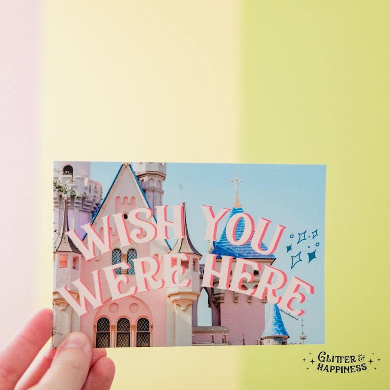 Pode incluir: Um cart&atilde;o postal com um castelo rosa e azul no fundo e o texto "Wish you were here" em letras rosa e azuis com um efeito brilhante.