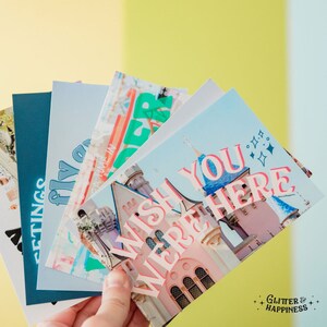 Pode incluir: Um conjunto de cinco cart&otilde;es postais coloridos com um castelo de conto de fadas e o texto "Wish You Were Here". Os cart&otilde;es postais s&atilde;o impressos em papel texturizado com um esquema de cores azul e rosa.