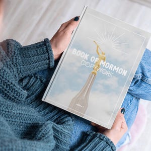 May include: A person is holding a book titled "Book of Mormon Copywork, Volume 1". The cover features a golden statue of an angel with a trumpet against a blue sky background.