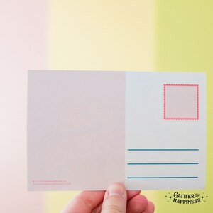 Pode incluir: Um cart&atilde;o postal branco em branco com um contorno de selo rosa e linhas azuis para escrever. O cart&atilde;o postal tem o texto "@glitterandhappiness glitterandhappiness.com" e "Glitter & Happiness" no canto inferior direito.