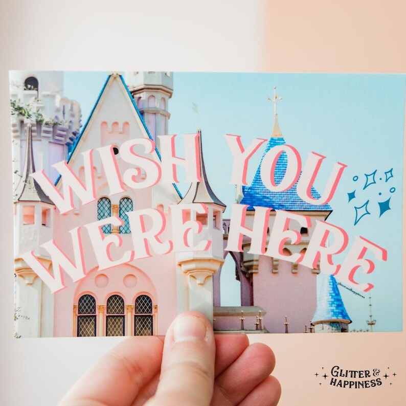 Pode incluir: Um castelo rosa e azul com o texto "Wish you were here" em letras brancas com contorno rosa. O texto &eacute; sobreposto &agrave; imagem do castelo. A imagem tem um fundo de c&eacute;u azul com brilhos brancos.