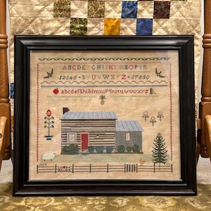 May include: A framed cross-stitch embroidery depicting a small, gray, wooden cabin with a red door, surrounded by a fence and a small pine tree. The embroidery includes a red apple, a pineapple, and a bird. The top of the frame includes a cross-stitch alphabet and numbers.
