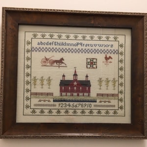 May include: Framed cross-stitch embroidery featuring a red barn with a white star, a horse-drawn carriage, a rooster weather vane, and a quilt block. The embroidery also includes the alphabet, numbers 1-10, and a border of green leaves.