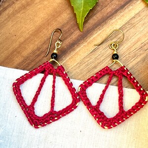 May include: A pair of red crocheted earrings with gold accents and black beads. The earrings are shaped like a diamond with a geometric design.