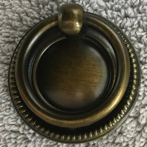 May include: A round, antique brass cabinet knob with a ring pull. The knob has a decorative, beaded edge.