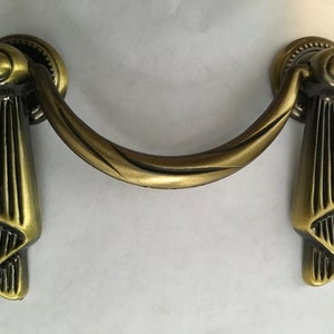 以下が含まれることがあります： 装飾的なスクロールデザインのアンティークゴールド調の引き出しプル。プルには、丸いノブが付いた2つの装飾的な端があります。