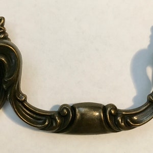 May include: Antique brass-toned drawer pull with an ornate design. The pull features an oval backplate with a beaded border and a decorative handle. The top of the backplate has a floral and ball design. A classic hardware piece.