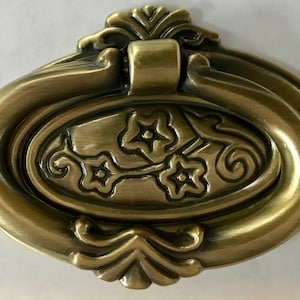 Puede incluir: Un tirador de armario de latón antiguo con un diseño floral. El tirador tiene un asa de anillo y detalles ornamentados.
