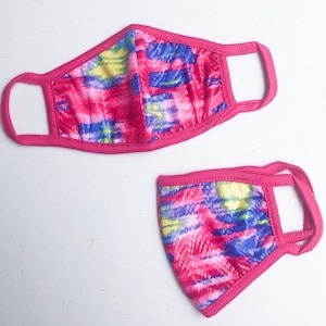 May include: Two pink face masks with a colorful abstract pattern. The masks are made of fabric and have elastic ear loops.