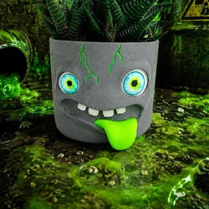 Pode incluir: Um vaso cinza com rosto de monstro com uma língua verde e detalhes verdes. Contém uma planta suculenta verde. O fundo apresenta um tema de resíduos tóxicos com líquido verde e um símbolo de perigo.