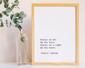 May include: A framed print with a quote by Khalil Gibran: "Beauty is not in the face; beauty is a light in the heart." The print is on a white background with black text and is framed in a light wood frame. A sprig of eucalyptus is in a clear glass vase on a white surface.