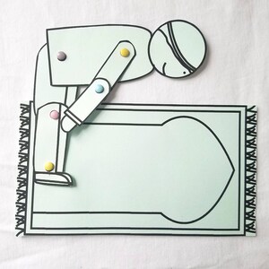 May include: A paper craft of a person kneeling on a prayer rug. The person is wearing a white head covering and has a black eye patch. The prayer rug is light blue with a black border and a black and white design in the centre.