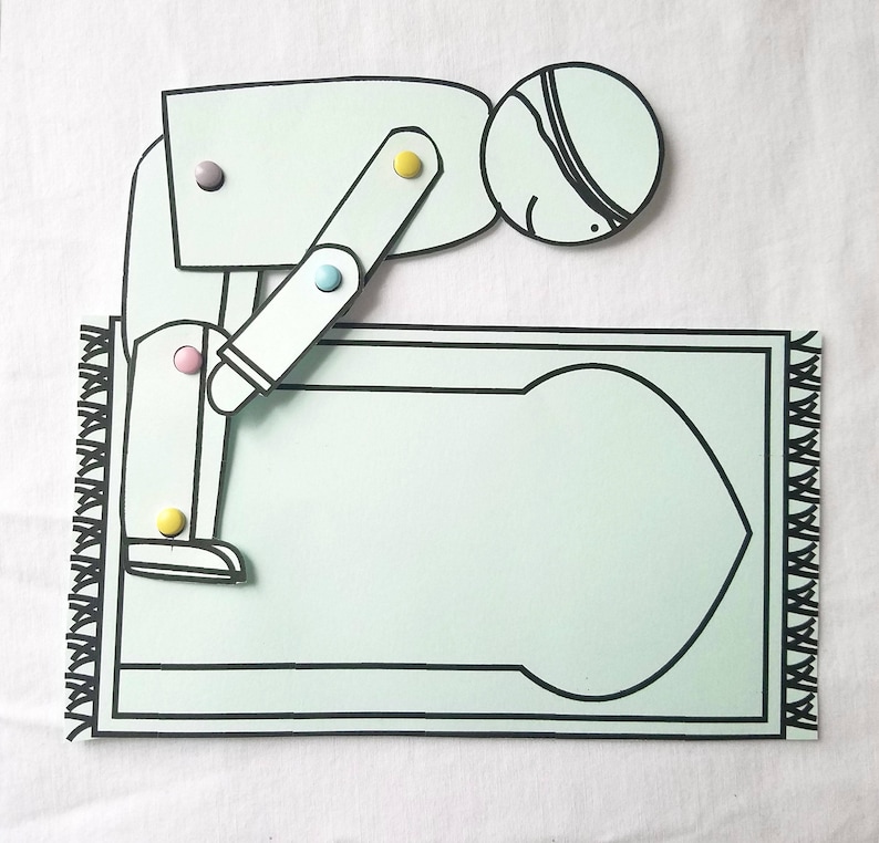 May include: A paper craft of a person kneeling on a prayer rug. The person is wearing a white head covering and has a black eye patch. The prayer rug is light blue with a black border and a black and white design in the centre.