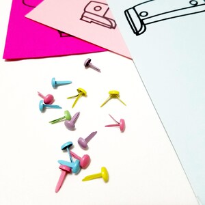 May include: Assortment of colourful metal brads in blue, pink, yellow, and purple. The brads are scattered on a white surface.