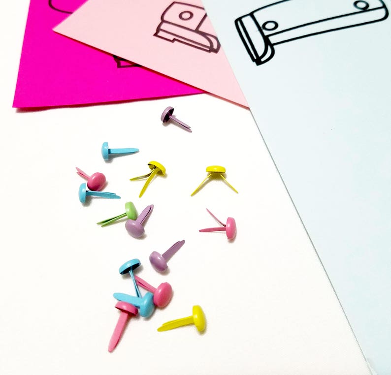 May include: Assortment of colourful metal brads in blue, pink, yellow, and purple. The brads are scattered on a white surface.