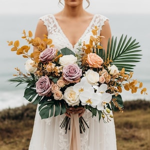 May include: A bridal bouquet featuring a variety of flowers in shades of white, peach, and lavender. The arrangement includes roses, orchids, and greenery, held by a person wearing a white lace dress. The background shows the ocean.