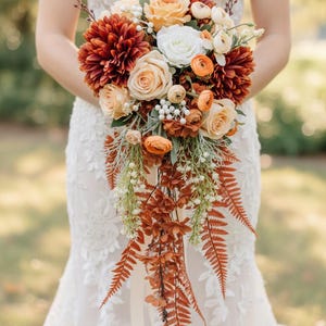 May include: A cascading bridal bouquet featuring a mix of flowers in shades of orange, peach, and white. The bouquet includes dahlias, roses, and other floral elements, with trailing ferns and greenery. The bouquet is held by a person wearing a white lace dress.