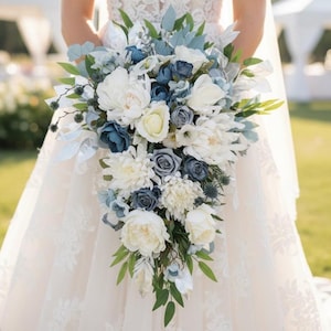Op de afbeelding: Een waterval bruidsboeket met witte pioenrozen, blauwe rozen en groen. Het arrangement bevat lichtblauwe en zilveren accenten, waardoor een zachte, elegante uitstraling ontstaat. Het boeket wordt vastgehouden tegen een witte kanten trouwjurk.