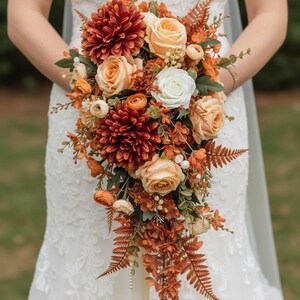 May include: A cascading bridal bouquet featuring a mix of orange, peach, and burgundy flowers. The bouquet includes roses, dahlias, and ferns, with a white rose accent. The flowers are arranged with greenery and a ribbon.