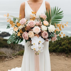 May include: A bridal bouquet featuring a variety of flowers in shades of pink, peach, and white, including roses and orchids. The bouquet is accented with green foliage and gold-toned leaves, tied with a blush ribbon.