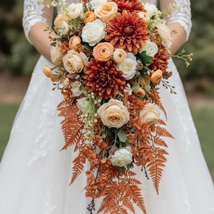 May include: A cascading bridal bouquet featuring a mix of flowers in shades of white, peach, and rust. The bouquet includes roses, dahlias, and ferns, creating a natural, elegant look. The bouquet is held in front of a white dress.