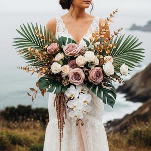 May include: A large bridal bouquet featuring a variety of flowers in shades of pink, white, and mauve, accented with green palm leaves and gold-toned foliage. The bouquet includes roses, orchids, and other floral elements, creating a lush and elegant arrangement.