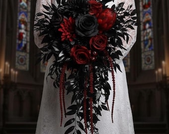 May include: A cascading bridal bouquet featuring black and deep red flowers. The arrangement includes roses, dahlias, and other blooms, accented with black foliage and long, red beaded strands. The bouquet is held against a white dress.