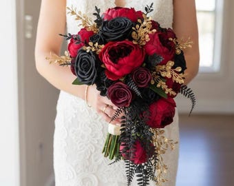 Puede incluir: Un ramo de novia con rosas rojas y negras, peonías y otras flores, acentuado con follaje dorado y hojas negras en forma de helecho. El ramo se sostiene contra un vestido de encaje blanco.