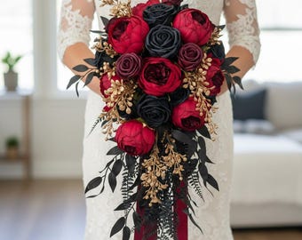 Puede incluir: Un ramo de novia en cascada con peonías rojas y rosas negras, acentuado con follaje dorado y negro. Cintas burdeos añaden un toque de elegancia. El ramo es sostenido por una persona que lleva un vestido de encaje blanco.