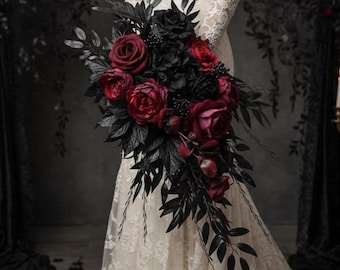 May include: A cascading bridal bouquet featuring deep red and black roses, peonies, and berries, accented with black leaves. The arrangement is held against a backdrop of a white lace dress, creating a dramatic contrast. The bouquet is a gothic or alternative wedding theme.