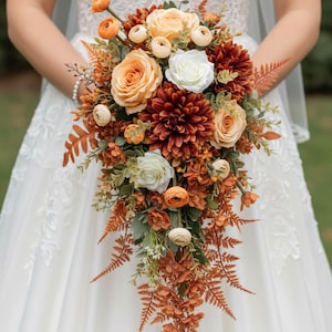 May include: A cascading bridal bouquet featuring orange, peach, and white roses, dahlias, and other flowers. The bouquet includes greenery and fern accents, creating a natural, elegant look. The flowers are held against a white dress.