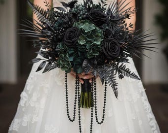 May include: A dark, gothic-style bridal bouquet featuring black roses, dark green hydrangeas, and various black foliage. The bouquet is accented with strands of black beads and wrapped in a black handle.